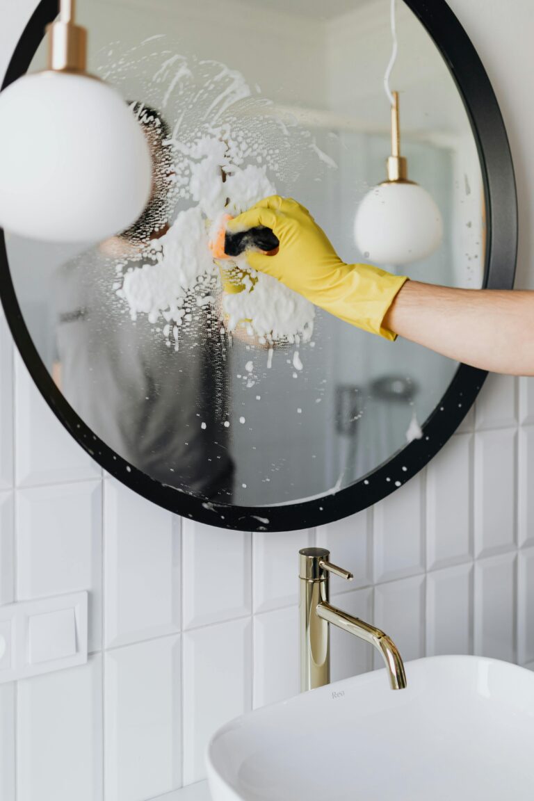 A person wearing a yellow glove cleans a bathroom mirror with foam spray.