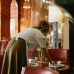 Home A woman is wiping a glass table in a cozy cafe with vintage decor and soft lighting.