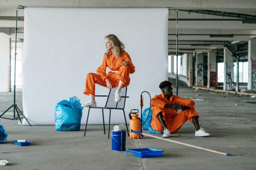 Two workers in orange uniforms pose in an industrial setting, surrounded by cleaning supplies.